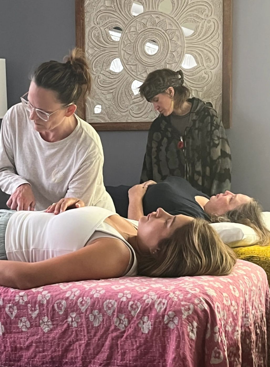 Two students giving Reiki to a client on a treatment table beneath a large mandala wall hanging