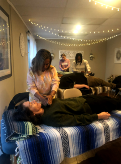 A practitioner holding her hands over a client resting on a treatment table, with other students working in the background